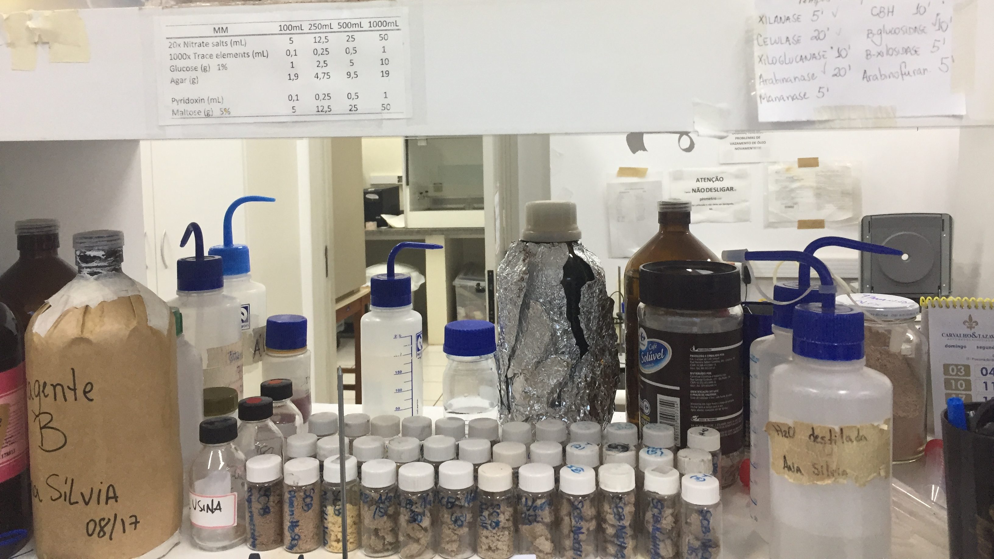 Plastic tubes and bottles of various sizes are lined up on a scientific lab bench. Some are filled with liquids, others are filled with brown chunks of cardboard-like biomass. Pieces of paper with recipes and other information are taped to the shelving.