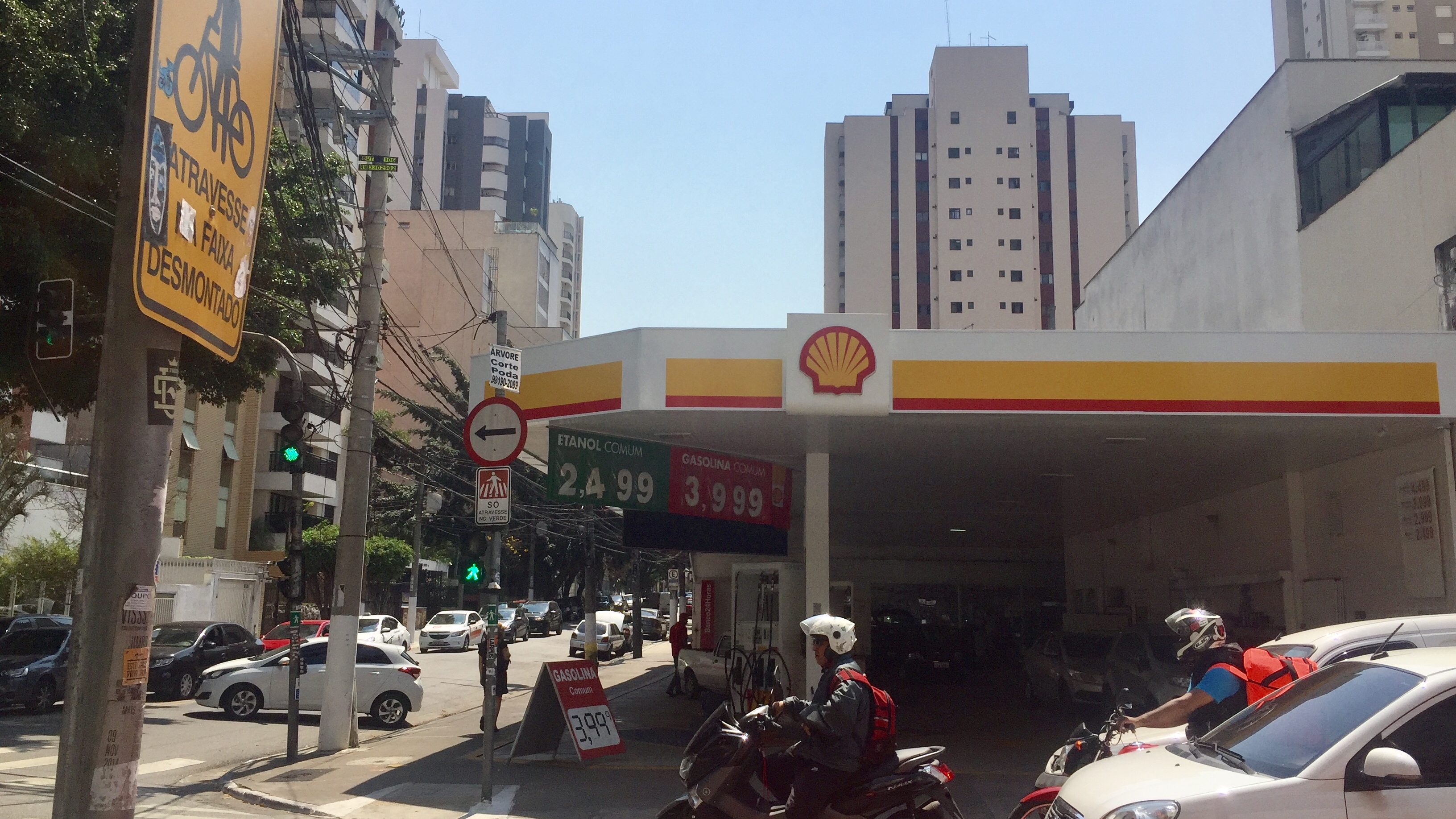 A Shell fuel station at a busy city intersection. Cars and motorcycles idle in front of it while waiting for the traffic light. Under the awning of the fuel station hangs two prices, one in green (2.499) and one in red (3.999).