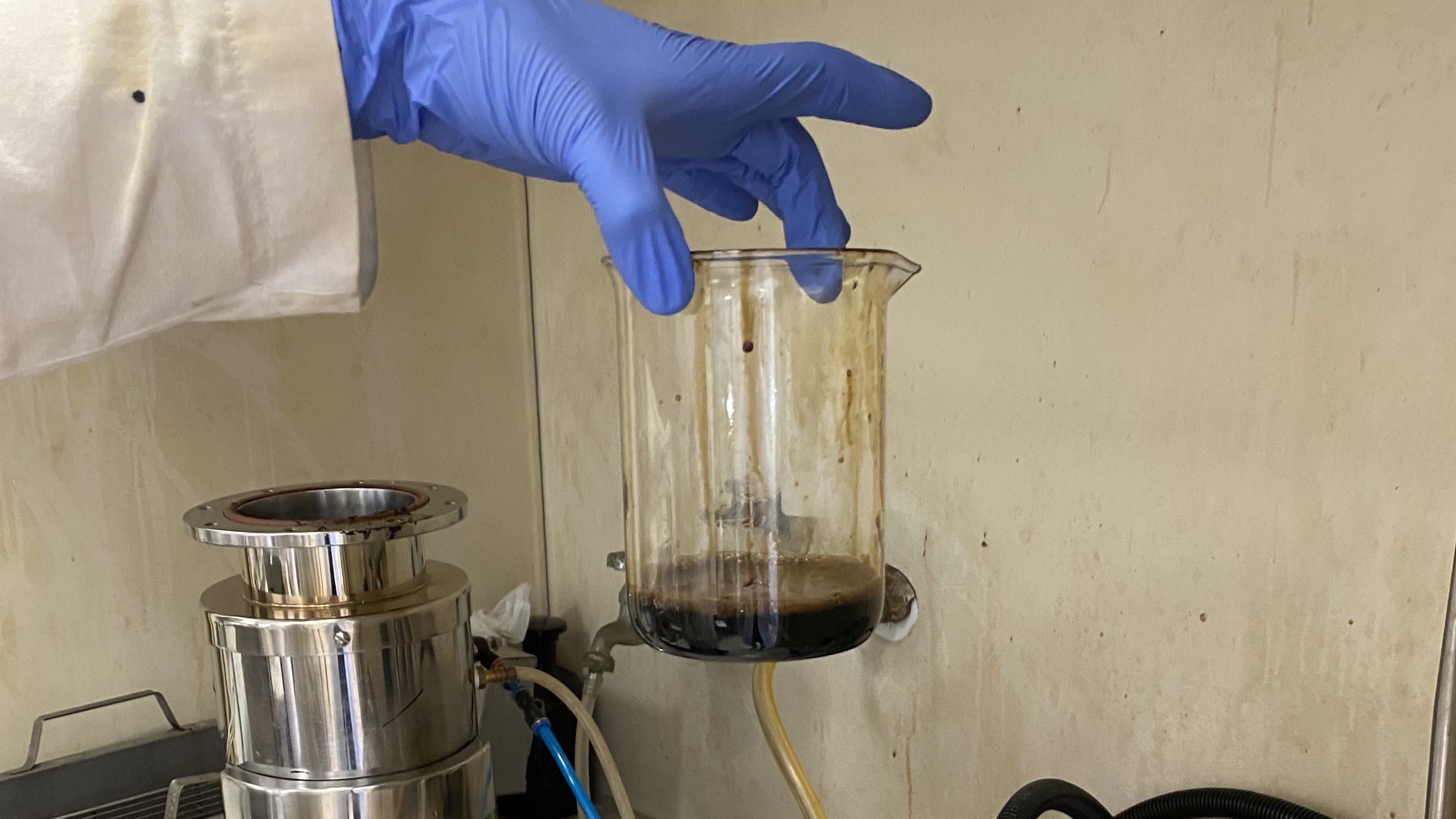 A hand wearing a blue nitrile glove holds a glass beaker from the top. At the bottom of the beaker is an inch or so of dark brown, slightly frothy liquid. In the background is the stained metal walls of a chemical hood. The top of a metal canister peaks into the frame on the lower left.
