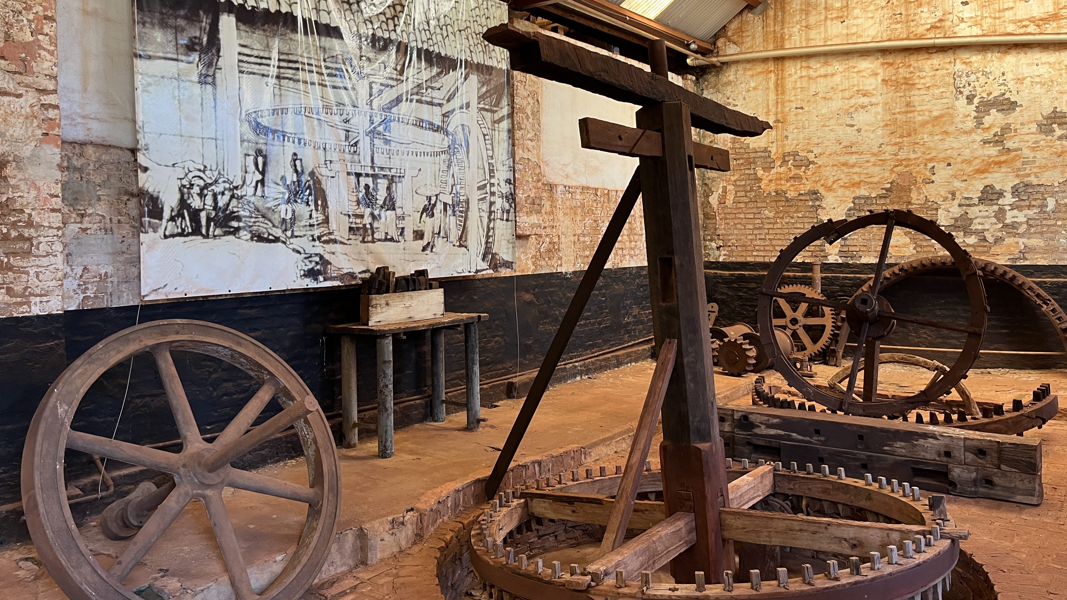 Rusted metal equipment, including wheel-like structures with gears, sits inside a brick room. On the wall is a large illustration of enslaved people of color using this equipment, alongside oxen, in an old sugar mill.