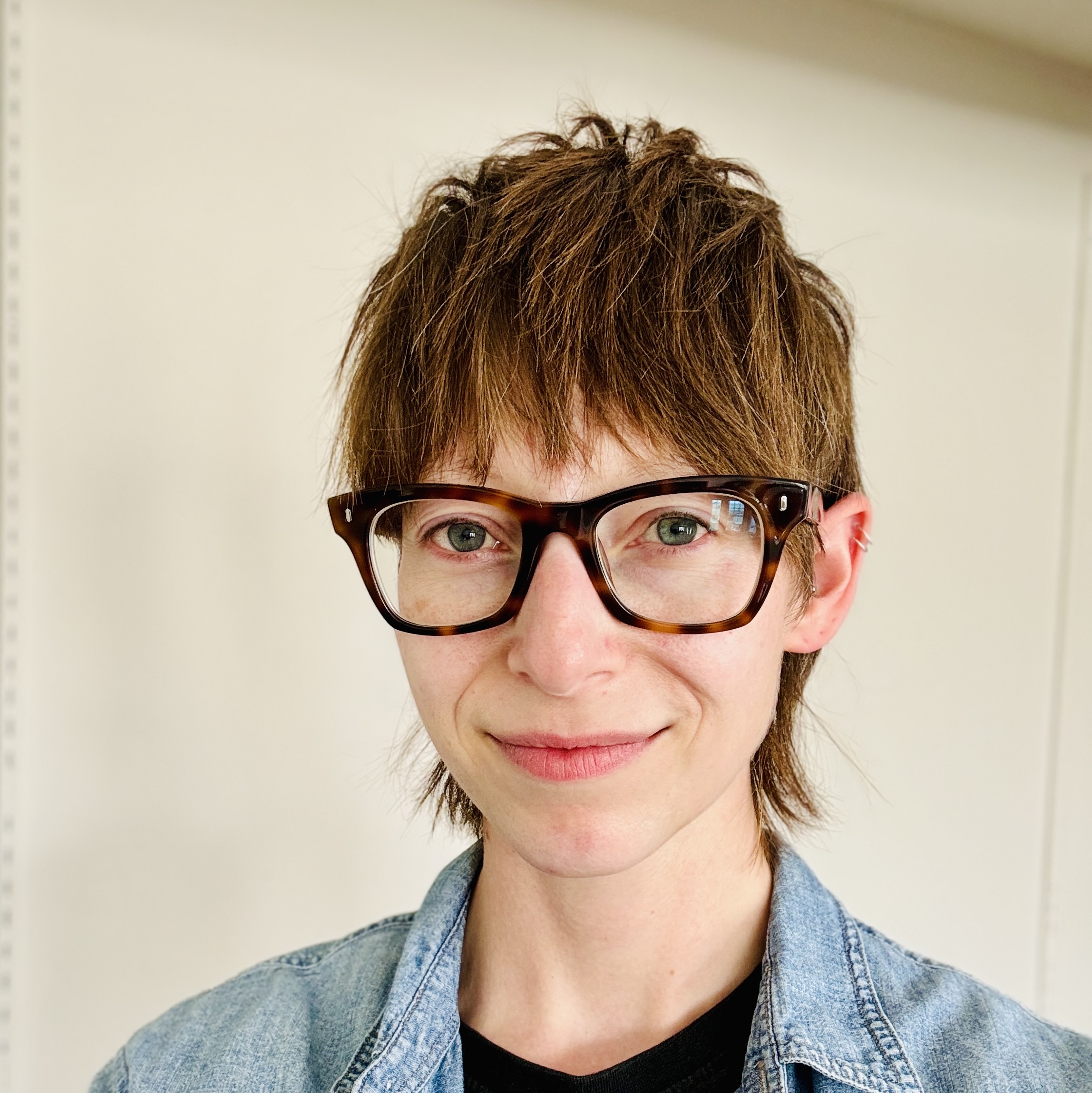 Headshot of a person with short brown hair and thick tortoise-shell glasses. They are smiling softly and where a denim button-up over a black shirt.